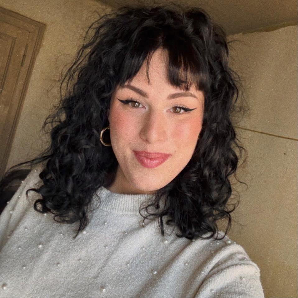 Selfie de Tamy en pull gris, cheveux bouclés, regard direct.
